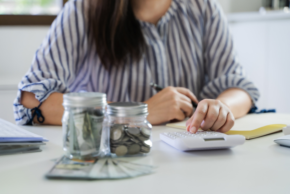 Woman siting at table, using a calculator with jars of coins and dollar bills around her.